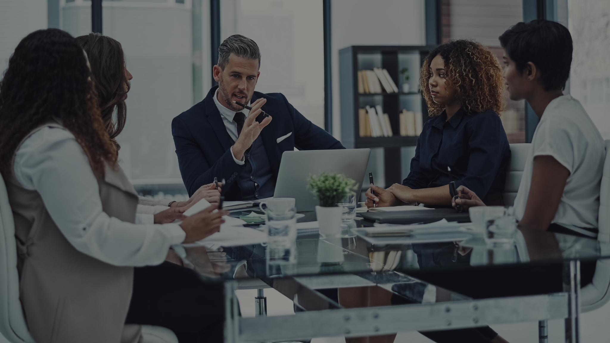 A corporate team engaged in a serious discussion around a glass meeting table in a modern office. The group, consisting of diverse professionals, listens to a suited man speaking and gesturing for emphasis. This scene underscores the importance of understanding U.S. regulations in a business context, as clear communication and compliance are essential.