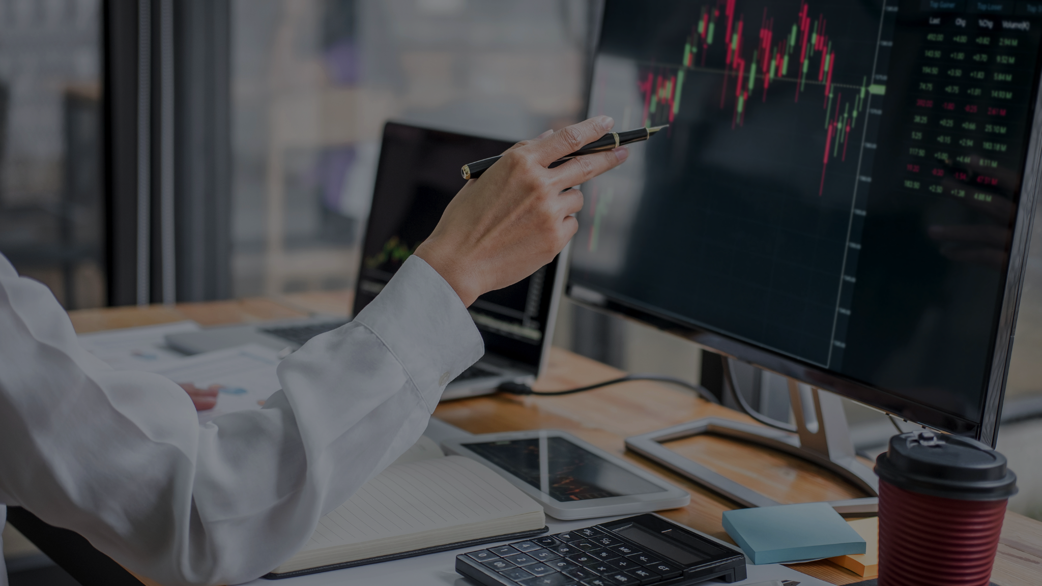 A professional pointing at a monitor displaying stock charts and market data in a modern office. The desk is equipped with tools like a calculator, tablet, and coffee cup, suggesting detailed analysis and planning. This image represents how international companies divide the U.S. market, focusing on data-driven strategies and market insights for effective decision-making.