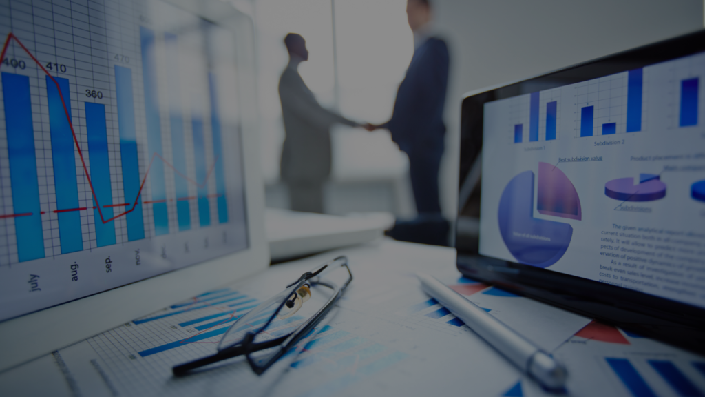 A workspace showcasing market entry analysis tools, with financial charts displayed on screens and papers. Glasses and a pen rest on the desk, while two professionals shake hands in the blurred background, emphasizing strategic collaboration.